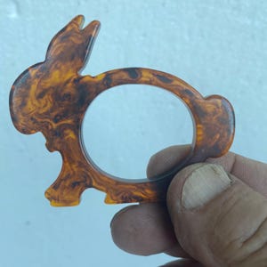 May include: A rabbit-shaped napkin ring crafted from a polished, marbled material featuring a swirling pattern of deep brown and amber tones. The design incorporates a circular opening in the centre, with the rabbit's ears, head, and tail clearly defined.