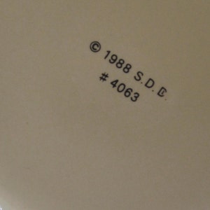 Pode incluir: Close-up de uma superf&iacute;cie creme com o texto "&copy; 1988 S.D.C. #4063" impresso em preto. A superf&iacute;cie parece fazer parte de um objeto maior, possivelmente cer&acirc;mica ou porcelana.