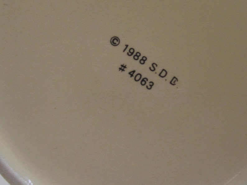 Pode incluir: Close-up de uma superf&iacute;cie creme com o texto "&copy; 1988 S.D.C. #4063" impresso em preto. A superf&iacute;cie parece fazer parte de um objeto maior, possivelmente cer&acirc;mica ou porcelana.