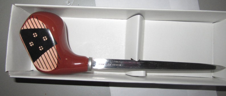 May include: A letter opener with a handle shaped like a brown golf club head featuring black and tan accents. The blade is made of silver-toned metal with the text STAINLESS STEEL etched onto the surface, resting inside a white box.