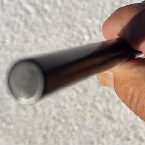 Puede incluir: Una varilla cilíndrica de madera de color marrón oscuro con un acabado liso y pulido. El objeto es recto y presenta una sección transversal circular en el extremo, mostrando una textura de veta de madera oscura, sólida y consistente.