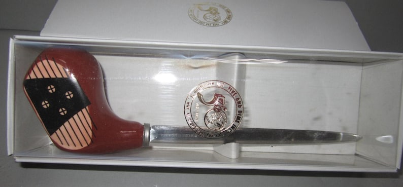 May include: A letter opener with a handle shaped like a brown golf club head featuring black and tan striped details. The metal blade is attached to a circular emblem that reads F.W. WOOLWORTH CO. FIVE AND DIME INC. It is presented in a white box.