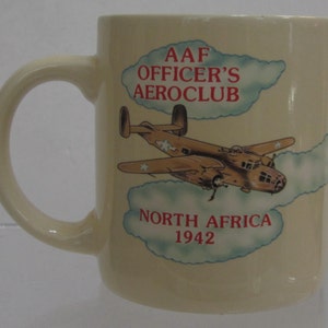 Pode incluir: Caneca de cer&acirc;mica creme com uma ilustra&ccedil;&atilde;o de avi&atilde;o vintage. A caneca tem o texto "AAF OFFICER'S AEROCLUB NORTH AFRICA 1942" em letras vermelhas. O design inclui um avi&atilde;o castanho com estrelas brancas.