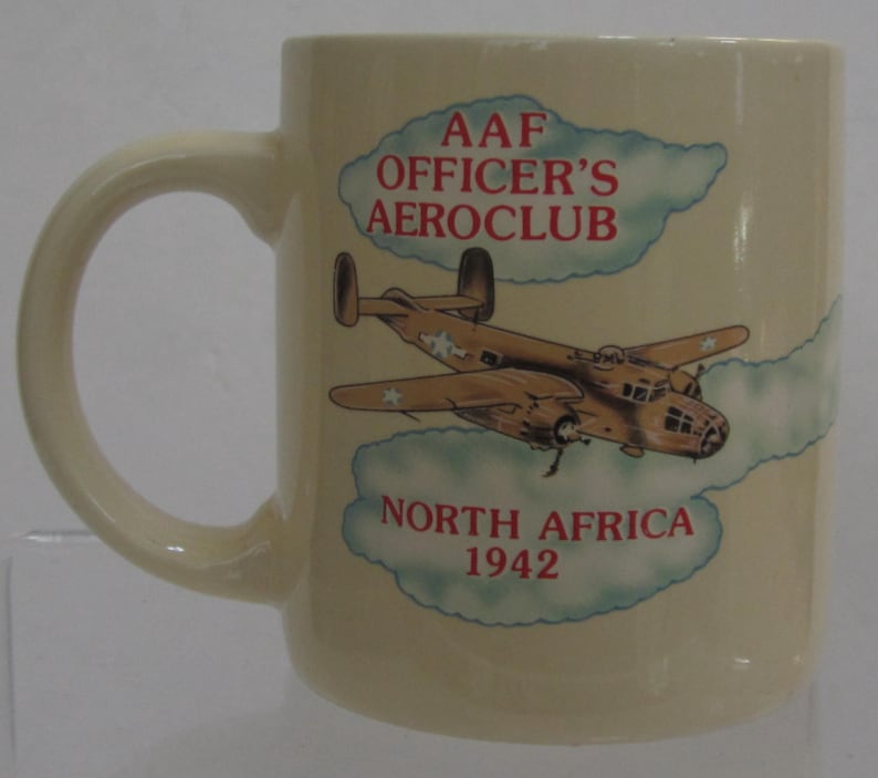 Pode incluir: Caneca de cer&acirc;mica creme com uma ilustra&ccedil;&atilde;o de avi&atilde;o vintage. A caneca tem o texto "AAF OFFICER'S AEROCLUB NORTH AFRICA 1942" em letras vermelhas. O design inclui um avi&atilde;o castanho com estrelas brancas.
