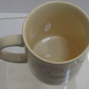 Pode incluir: Caneca de cer&acirc;mica creme com uma al&ccedil;a curva. O interior &eacute; um tom de creme mais claro. A caneca tem texto e um gr&aacute;fico na lateral. A caneca foi projetada para bebidas quentes.
