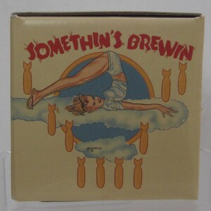 Pode incluir: Caixa bege com a inscri&ccedil;&atilde;o vermelha "SOMETHIN'S BREWIN". A caixa apresenta uma ilustra&ccedil;&atilde;o vintage de uma mulher em pose de ponte sobre uma nuvem, rodeada por bombas de desenho animado. A obra de arte tem um estilo retro pin-up.