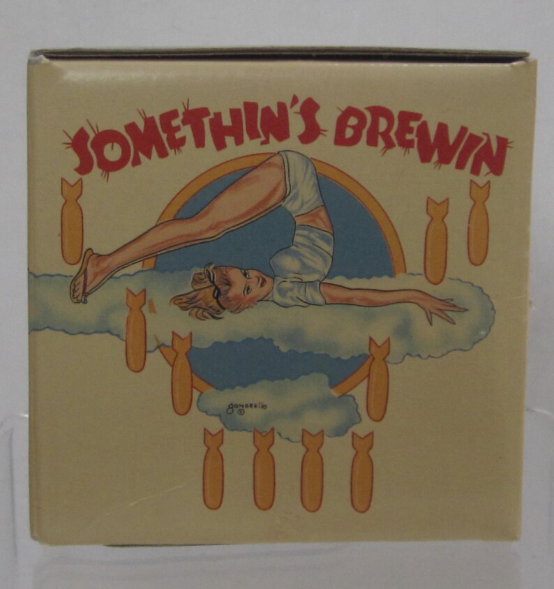 Pode incluir: Caixa bege com a inscri&ccedil;&atilde;o vermelha "SOMETHIN'S BREWIN". A caixa apresenta uma ilustra&ccedil;&atilde;o vintage de uma mulher em pose de ponte sobre uma nuvem, rodeada por bombas de desenho animado. A obra de arte tem um estilo retro pin-up.