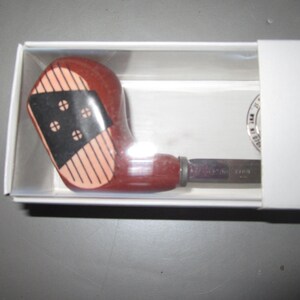 May include: A bottle opener with a handle shaped like a vintage wooden golf club head. The handle is reddish-brown with a black face insert featuring four white circular markings and vertical lines, encased in a white cardboard box.