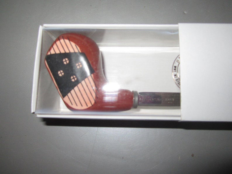 May include: A bottle opener with a handle shaped like a vintage wooden golf club head. The handle is reddish-brown with a black face insert featuring four white circular markings and vertical lines, encased in a white cardboard box.
