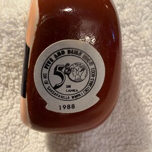 May include: A smooth, dark brown ceramic object featuring a circular white sticker on its base. The sticker displays the text Five and Dime, Div of Sarsaparilla, Sri Lanka, and the year 1988, along with a graphic of a profile portrait.