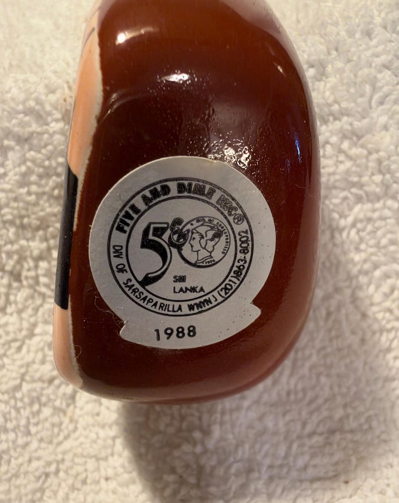 May include: A smooth, dark brown ceramic object featuring a circular white sticker on its base. The sticker displays the text Five and Dime, Div of Sarsaparilla, Sri Lanka, and the year 1988, along with a graphic of a profile portrait.