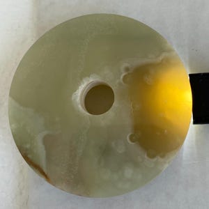 Pode incluir: Um disco de pedra circular de cor verde pálido com um orifício central, apresentando inclusões brancas translúcidas e nubladas com uma superfície lisa e polida. Uma luz amarela brilhante ilumina um lado, realçando a textura mineral.