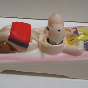 Puede incluir: Un juego de salero y pimentero de cerámica. El juego representa una figura reclinada con una toalla roja, una mujer rubia y dos saleros y pimenteros. La base es blanca con un ribete rosa.