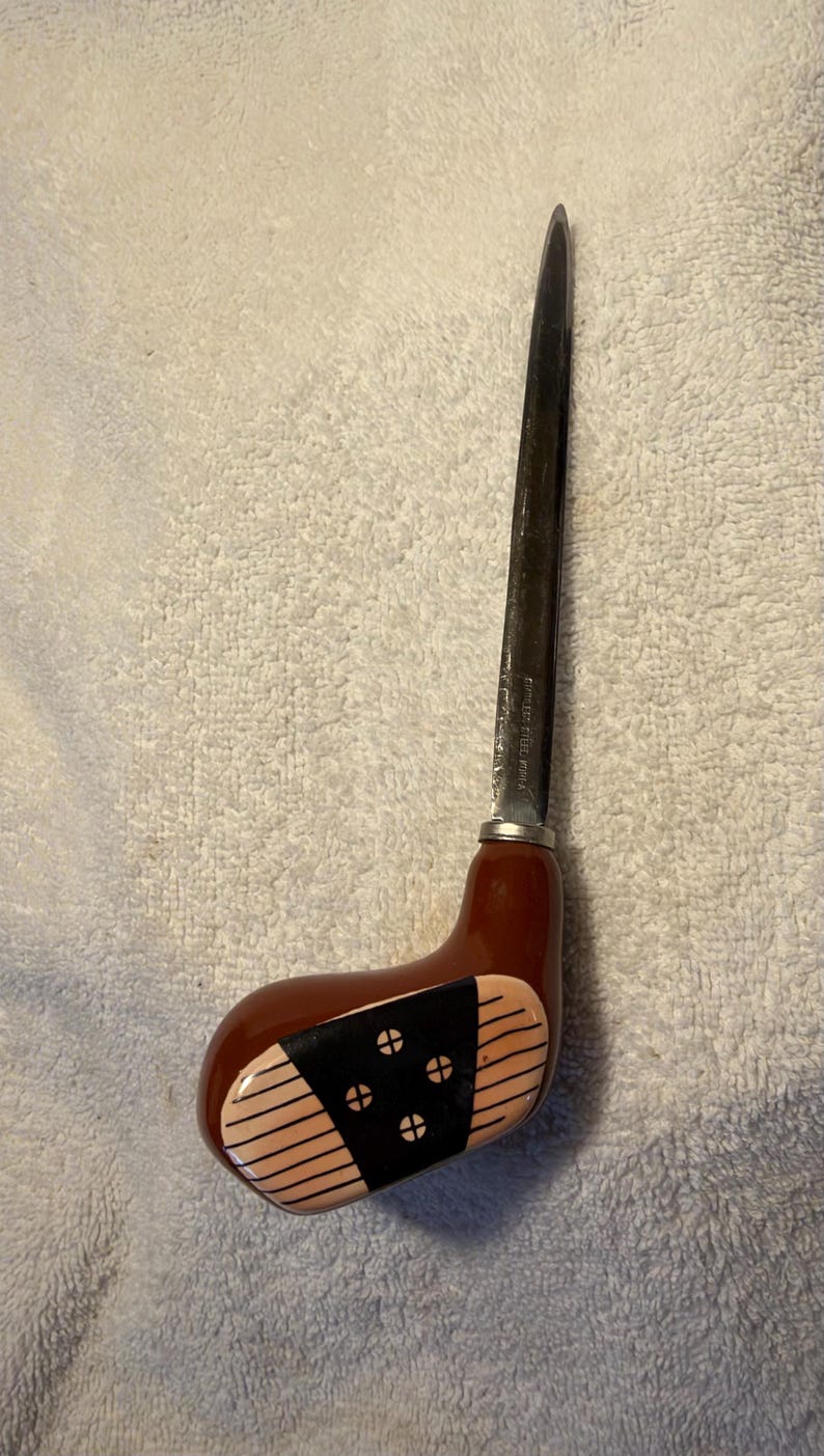 May include: A letter opener featuring a metal blade attached to a handle shaped like a brown golf club head. The handle has a black center panel with four circular accents and thin horizontal lines on the light tan sides. The blade is marked Stainless Steel Korea.