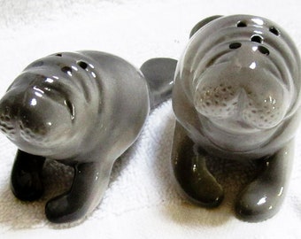 May include: A pair of ceramic manatee salt and pepper shakers. The gray manatees are in a sitting position, with small holes on top for dispensing. The manatees have a glossy finish and detailed features.