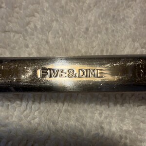 May include: A metallic, rectangular object with a polished, reflective surface. The words FIVE & DIME are embossed in a clear, sans-serif font across the center of the metal bar, showing light scratches and signs of wear.