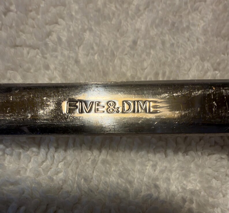 May include: A metallic, rectangular object with a polished, reflective surface. The words FIVE & DIME are embossed in a clear, sans-serif font across the center of the metal bar, showing light scratches and signs of wear.