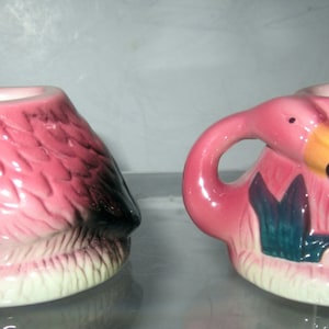 Może przedstawiać: Dwa ceramiczne kubki w kształcie flamingów. Kubki są różowe z czarnymi, żółtymi i turkusowymi akcentami. Głowa flaminga tworzy uchwyt, a korpus to kubek. Kubki są zaprojektowane ze wzorem tekstury piór.