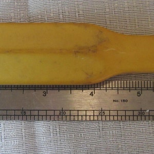 Puede incluir: Una herramienta de plástico amarillo vintage con una cabeza plana y rectangular y un mango cónico. La herramienta mide aproximadamente 18 cm de largo. Se coloca una regla de metal junto a la herramienta.