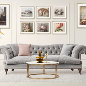 May include: A grey tufted sofa with a pink and a grey pillow, positioned before a white wall featuring eight framed art prints. A round coffee table with a white top and gold base holds a brown glass vase and a gold candle holder.