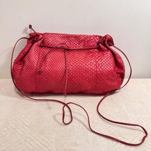 May include: A vibrant red handbag featuring a textured snakeskin pattern. The bag has a soft, slouchy silhouette with a drawstring closure mechanism and a thin, matching red shoulder strap that extends from the sides of the main compartment.