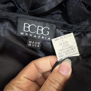 May include: A close-up of a black garment with the brand label "BCBG MAXAZRIA" and a tag that reads "100% POLYESTER" and "MADE IN U.S.A.". The garment appears to be made of a shiny, silky material.