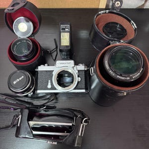 May include: A vintage Nikkormat camera with multiple lenses and accessories. The camera is silver and black, with a flash unit attached. Several lenses are visible, along with their cases and a Nikon lens cap. A black camera bag is also present.