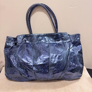 May include: A dark blue handbag featuring a glossy, textured snakeskin pattern. The bag has two rounded top handles, a gathered, pleated front design, and two small circular decorative buttons positioned near the top edge of the main compartment.