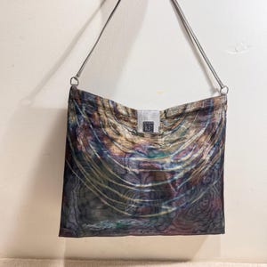 Peut inclure: Un sac à bandoulière rectangulaire en tissu translucide et irisé, orné de motifs tourbillonnants dans des tons bronze, or, vert et violet. Le sac comprend une fine bandoulière argentée et un fermoir central en métal argenté avec le texte B.V.G.T.