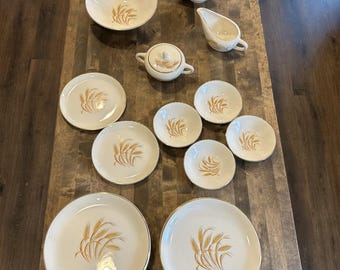 May include: A collection of vintage white ceramic dishware with a wheat stalk design in gold. The set includes plates, bowls, saucers, a sugar bowl, and gravy boats. The edges of the plates and bowls have a gold trim.