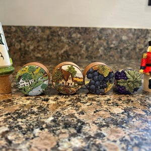 May include: A selection of decorative bottle stoppers with wooden bases. The stoppers showcase diverse designs, including a rural scene, a house, and a cluster of grapes. These stoppers are designed to fit into wine bottles.