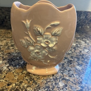 Puede incluir: Un jarrón de cerámica rosa pálido con borde festoneado y un diseño floral en relieve de color blanco que muestra dos flores prominentes y varias hojas. El jarrón tiene un acabado liso y brillante con un cuerpo ancho y redondeado.
