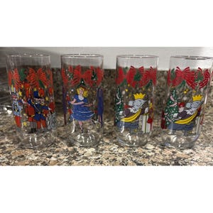 May include: Four clear glass tumblers with Christmas-themed illustrations. Each glass features a different design, including a toy soldier, a ballerina, and a mouse king. The rims are decorated with red bows and green foliage.