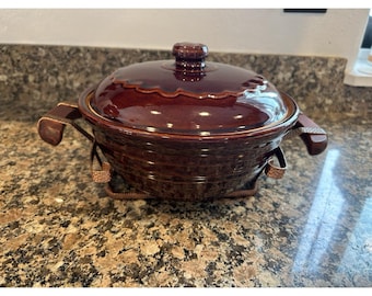 Puede incluir: Una fuente de cerámica marrón con tapa y asas. El plato tiene un acabado brillante y un borde decorativo. Se encuentra en un soporte de metal con detalles tejidos. Una pieza clásica para servir y cocinar.