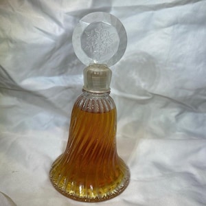 May include: A vintage perfume bottle with a clear, faceted stopper featuring a snowflake design. The bell-shaped bottle has a spiral design and is filled with amber-colored liquid. The bottle is made of glass.