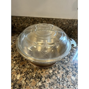 Puede incluir: Una olla redonda de metal con tapa de vidrio transparente. La tapa presenta un diseño central grabado de un molino de viento y herramientas cruzadas. La olla tiene dos asas laterales con acabado texturizado y una superficie interior lisa.