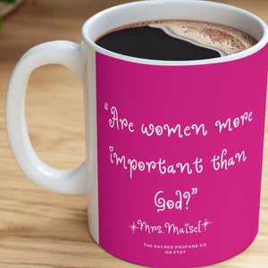 May include: A pink ceramic coffee mug with a white handle. The mug has a quote from the show "The Marvelous Mrs. Maisel" printed on it in white text: "Are women more important than God?"  Mrs. Maisel. The Sacred Profane Co. on Etsy.