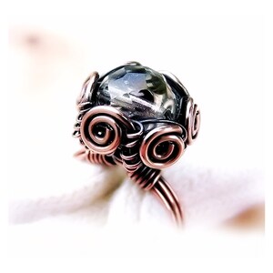 May include: A copper wire ring with a clear, faceted gemstone set in the centre. The wire is wrapped around the stone in a decorative pattern.