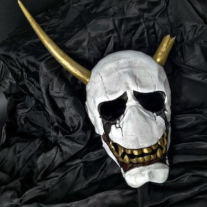 Può includere: Una maschera a teschio bianca con orbite nere e vernice gocciolante. La maschera ha corna e denti color oro. La maschera è esposta su un tessuto nero strutturato.