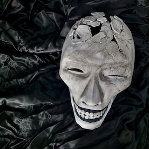 Può includere: Una maschera grigia e screpolata con un ampio sorriso dentato e occhi chiusi. La parte superiore della maschera è rotta, rivelando l'interno. La maschera è posta su uno sfondo scuro e strutturato.