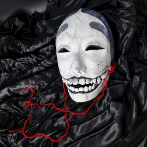 Può includere: Una maschera bianca con fessure nere per gli occhi e un ampio sorriso dentato. La maschera ha accenti blu sulla fronte ed è collegata con un filo rosso. La maschera poggia su un tessuto nero drappeggiato.