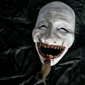 Può includere: Una maschera bianca sorridente con denti affilati e dettagli rossi. La maschera ha una lunga barba marrone ed è posta su uno sfondo scuro e strutturato. La maschera è probabilmente per costumi o decorazioni.
