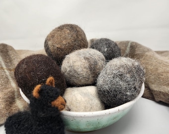 Puede incluir: Un bol lleno de bolas de lana de fieltro marrón, gris y blanco. El bol es azul claro con motas blancas. Una pequeña figura de alpaca negra está en primer plano.