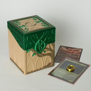 May include: A rectangular deck box featuring a tan base with embossed arch patterns and a metallic green lid adorned with intricate leaf and vine motifs. Beside the box lie two rectangular trading cards and a small gold-coloured metal ring.