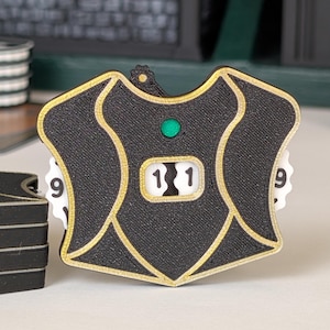 May include: A black and gold shield-shaped game counter featuring a central display showing the number 11. The device includes two white rotating dials on the sides with visible numbers, and a small green circular accent positioned above the display.
