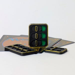 Könnte beinhalten: Ein vertikaler Spielzähler in Schwarz und Gold mit drei Schiebereglern, die die Zahl Null anzeigen, begleitet von zwei kleineren horizontalen Spielzählern, die auf mehreren Magic: The Gathering-Spielkarten liegen.