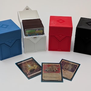 Könnte beinhalten: Vier Kartenaufbewahrungsboxen in Blau, Weiß, Rot und Schwarz, mit einem Diamantmuster auf dem Deckel. Die weiße Box ist geöffnet und zeigt einen Stapel Karten. Drei Sammelkarten werden vor den Boxen angezeigt.