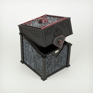 Puede incluir: Una caja negra con tapa abatible, que presenta una base con patrón de ladrillos grises y un panel superior decorado con un relieve de calavera roja. La tapa tiene un emblema de calavera roja más pequeño en el borde frontal y volutas negras.