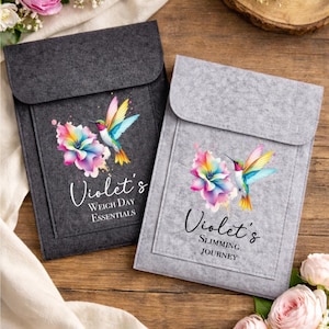 May include: Two felt pouches, one dark grey and one light grey, featuring a vibrant hummingbird and floral design. The dark grey pouch reads "Violet's Weigh Day Essentials." The light grey pouch reads "Violet's Slimming Journey."