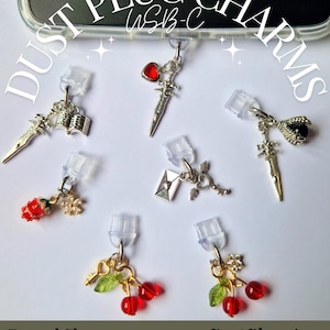 May include: Several silver-coloured dust plug charms with diverse designs, such as a sword, a heart-shaped gem, a book, a rose, a key, and cherries. The charms are connected to clear USB-C dust plugs. The image text says "Dust Plug Charms" and "Enamel Charm USB-C".